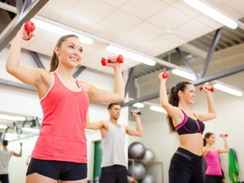 Vier Personen heben Gewichte in einem Fitnessstudio. Zwei Frauen tragen Sporttops, ein Mann hat ein T-Shirt an.