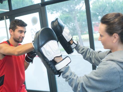 Ein Mann im roten Trikot schlägt auf gepolsterte Boxhandschuhe, die eine Frau mit grauem Hoodie hält.