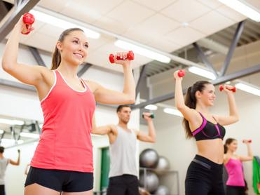 Vier Personen heben Gewichte in einem Fitnessstudio. Zwei Frauen tragen Sporttops, ein Mann hat ein T-Shirt an.