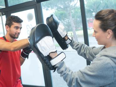 Ein Mann im roten Trikot schlägt auf gepolsterte Boxhandschuhe, die eine Frau mit grauem Hoodie hält.