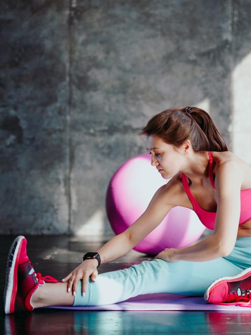 Eine Frau in Sportbekleidung dehnt ihren rechten Oberschenkel auf einer Yogamatte in einem Fitnessraum.