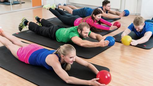 Fünf Personen liegen auf Gymnastikmatten und strecken sich nach bunten Fitnessbällen in einem Trainingsraum aus.