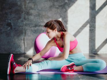 Eine Frau in Sportbekleidung dehnt ihren rechten Oberschenkel auf einer Yogamatte in einem Fitnessraum.