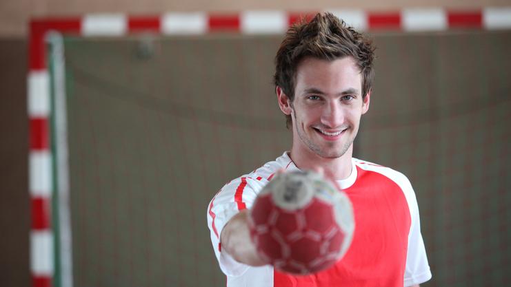 Junger Mann im roten Trikot hält einen Handball vor sich, lächelt in einer Sporthalle mit Tor im Hintergrund.