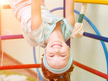 TG Kindersportschule Mädchen hängt kopfüber an einem Spielgerüst und lächelt, während sie rosa Ringe in den Händen hält.