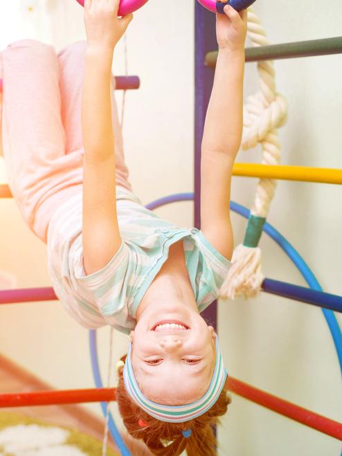 Mädchen hängt kopfüber an einem Spielgerüst und lächelt, während sie rosa Ringe in den Händen hält.