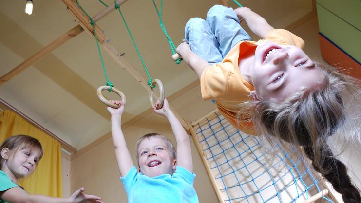 Drei Kinder spielen in einem Raum: Ein Junge hängt an Ringen, während zwei andere lachen und zuschauen.