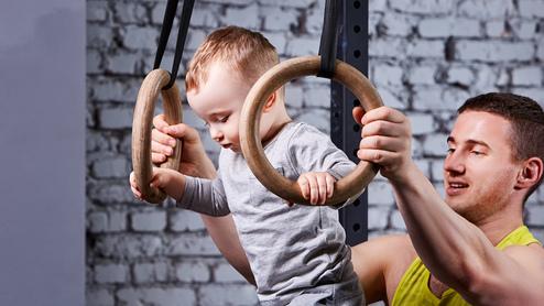 Ein Baby schwingt an Holzturnringen, während ein Erwachsener unterstützend daneben steht. Hintergrund mit Ziegelmauer.