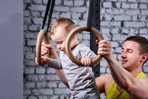 Ein Baby schwingt an Holzturnringen, während ein Erwachsener unterstützend daneben steht. Hintergrund mit Ziegelmauer.