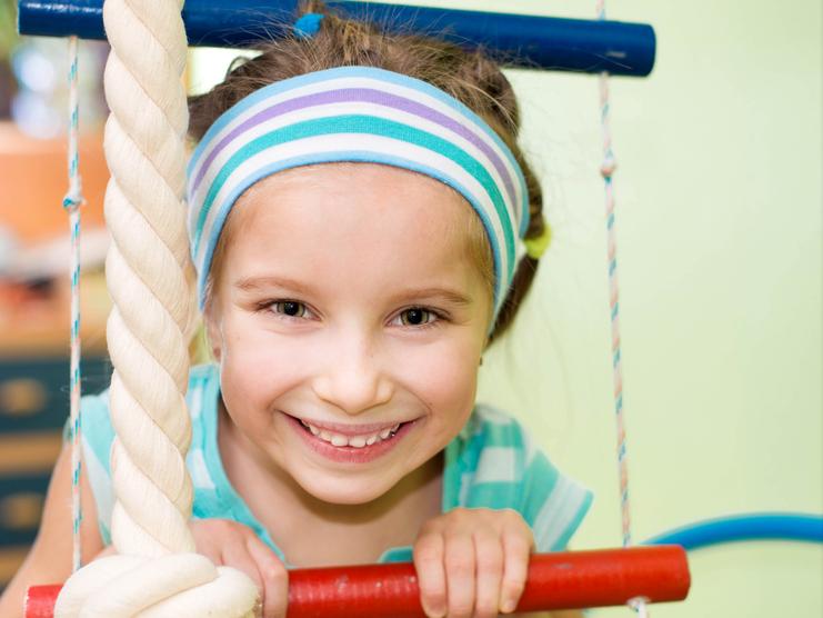 Lächelndes Mädchen mit blauer Kopfband und gestreifter Bluse hält sich an einem Klettergerüst fest.