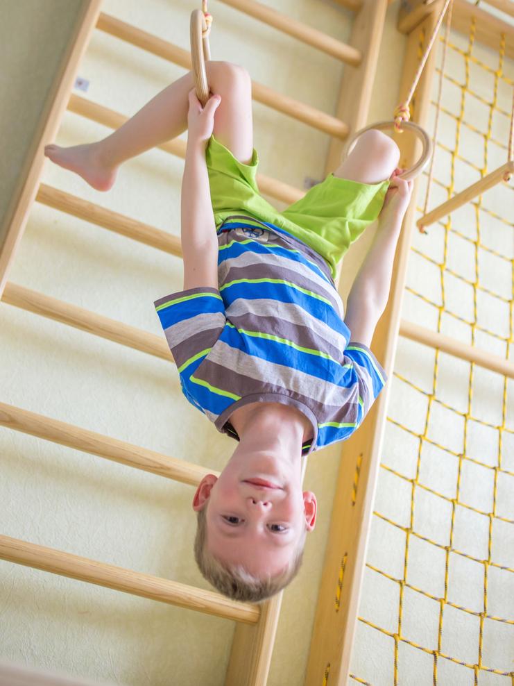 Junge hängt kopfüber an Turnringen vor einer Holzleiterspirale in einem hellen Raum.
