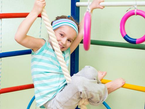 Ein Mädchen hängt lachend an einem Kletterseil in einer Turnhalle, umgeben von bunten Turngeräten.