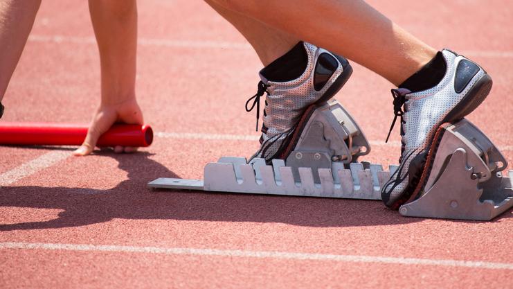 Startposition eines Sprintläufers mit Fuß auf einem Startblock auf einer roten Laufbahn, Hand hält einen Staffelstab.