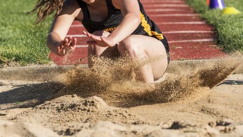 Athletin springt in einen Sandgruben, während Sand aufwirbelt. Grüße des Sprungbretts sind im Hintergrund sichtbar.