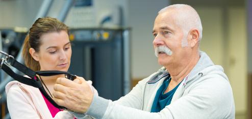 Ein älterer Mann mit Bart führt eine Übung mit einem Fitnessgerät unter Anleitung einer jungen Frau durch.