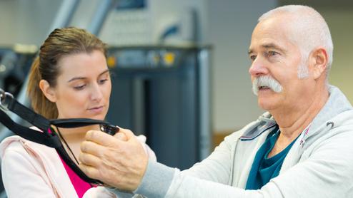 Ein älterer Mann mit Bart führt eine Übung mit einem Fitnessgerät unter Anleitung einer jungen Frau durch.