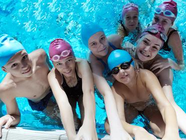 Gruppennahaufnahme von sieben Schwimmern im Wasser, lächelnd, mit Schwimmkappen und Brillen, vor einem blauen Hintergrund.