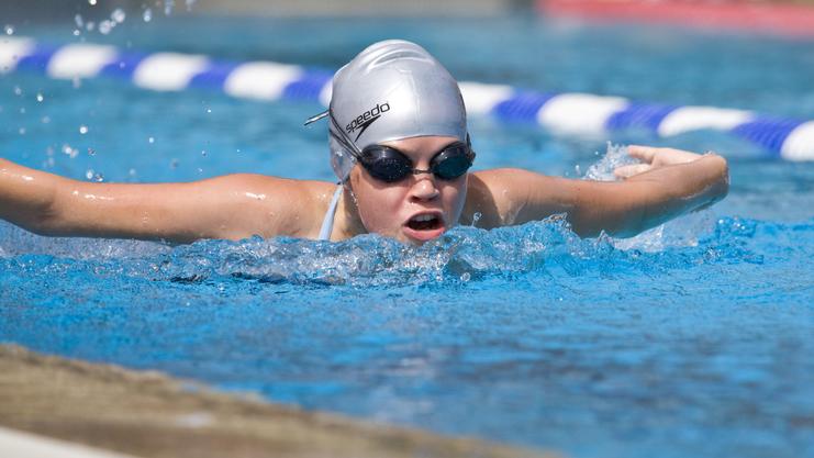 Junge Schwimmerin mit silberner Badekappe und Schwimmbrille in einem Wettkampfbecken, während sie einen Schlag ausführt.