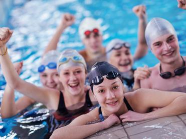 Sechs Jugendliche in Badekappen und Schwimmbrillen posieren fröhlich im Wasser, einige mit erhobenen Armen.