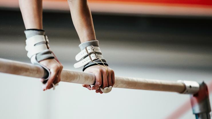 Nahaufnahme zweier Hände, die eine schmale Turnstange mit Griffpolstern halten, im Hintergrund eine Turnhalle.