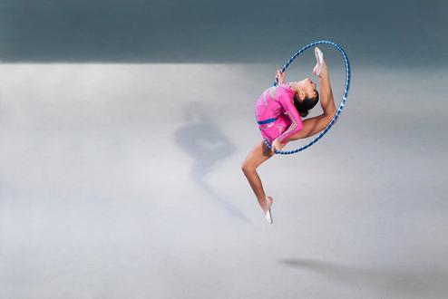 Mädchen in einem pinken Trikot springt mit einem Hula-Hoop-Reifen, während sie einen eleganten Tanzschritt ausführt.