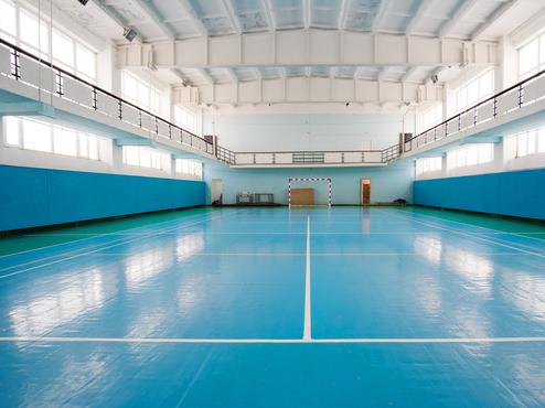 Leerer Sporthalle mit blauer Bodenfläche, seitlichen Wänden und Tor auf der gegenüberliegenden Seite.