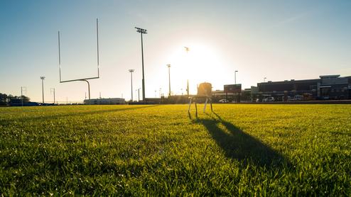 Sonnenuntergang auf einem Sportfeld mit einer einsamen Holzbank im Gras und Football-Tor im Hintergrund.